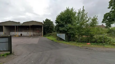 Google Maps A Google maps picture of the proposed site. It shows a wide road running through an open gate leading to barn-type buildings. There are trees and shrubs at the gate.