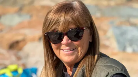 Tina in her 60s sat on Gorey beach. She is wearing a padded vest over a dark top. She has straight, shoulder‑length hair, and there is a colourful bag and a rocky surface in the background.