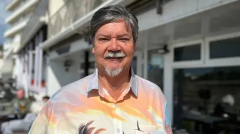Geoff Mayger is wearing a shirt with palm trees and flamingos on it, and is standing infront of his hotel. He has shirt dark hair and a grey goattee.