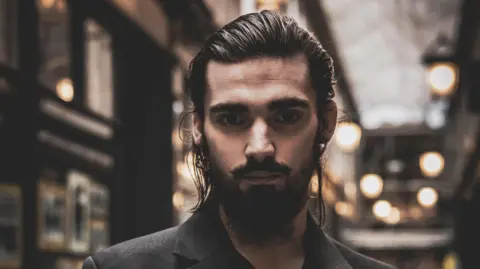 Tom Wilkins Theo, one of Tom's subjects, is pictured inside Cardiff's arcades. He has brown eyes and a brown beard, with long hair pulled back. Lights can be seen in the background but it's blurred. 