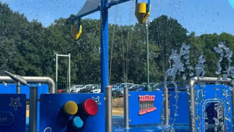 A splash park which has a blue floor and there are blue plastic panels propped up vertically by silver grey poles. There are jets spraying water into the air.