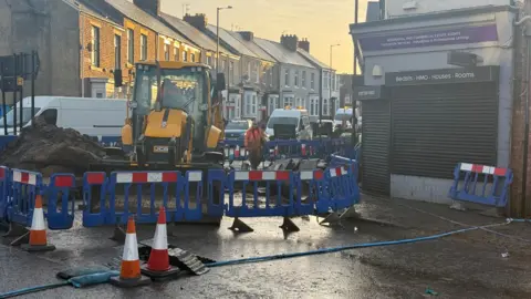 Workman carrying out roadworks to repair the damaged water main on Chester Road. A yellow JCB excavator is surrounded by blue plastic fencing. A pile of earth sits next to it. Traffic cones have been placed on the road and a thin, blue pipe stretches across it.