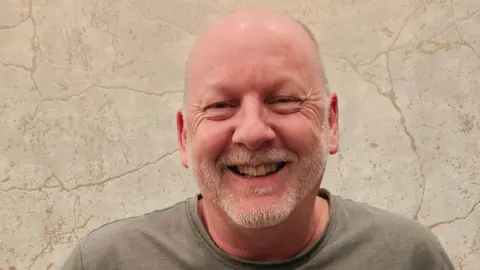 Chris Cramp A man wearing a grey t-shirt. He is stood in front of a stone wall.