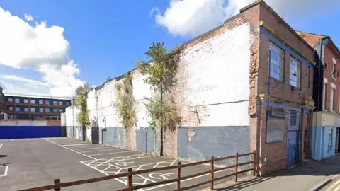 A scruffy looking building with exposed brick and plants growing out the side of it. There is a car park with disabled spaces and it is bright clear day with blue skies. 