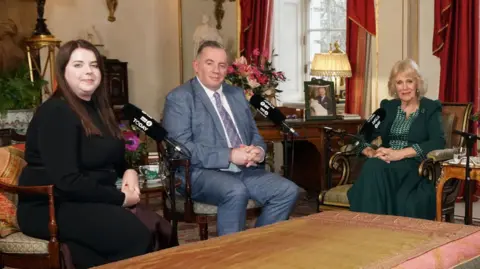 Neil Paton/Buckingham Palace/PA Wire Amy Hunt, a woman with brown hair wearing a black long sleeved dress, and John Hunt wearing a grey checked suit sit with Queen Camilla who wears a dark green outfit.  Baroness Theresa May and Emma Barnett  were also sat around a table in Clarence House but out of shot.
