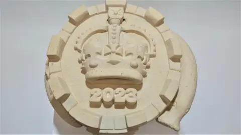 A circular piece of stone with a monarch's crown in the middle, 2023 written underneath it and a lamprey snaking around its sides.