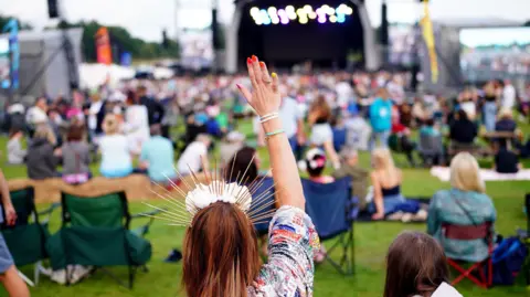 PA Media A woman with red hair and a sparkly headband holding her hand up, with a stage and crowd out of focus in the background