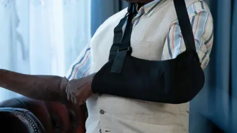 A close-up image of  man's left arm in a black sling. He is also wearing a white waistcoat and striped, short-sleeved shirt.