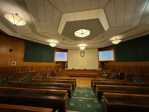 A large hexagonal room with rows of large green benches behind wooden desk. They are centred around a dais on the far wall beneath the Durham County logo