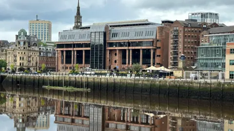 Newcastle Crown Court reflected in the River Tyne running in front of it. It is an imposing building made from smooth red stone with massive black windows and tall columns along its frontage.