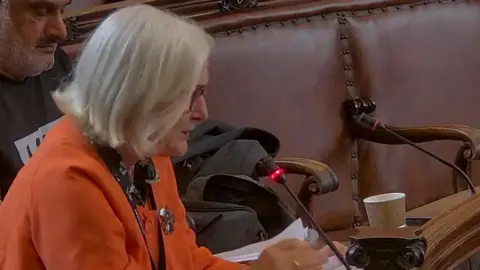 Wakefield Council Dodd has short, light-coloured hair is seated at a desk or bench and is speaking into a slim microphone. The microphone has a small red light illuminated, indicating that it is active. She is wearing an orange jacket over a darker top and has glasses on. In front of her, there are several sheets of paper or a document, which she appears to be reading from or referring to while speaking.