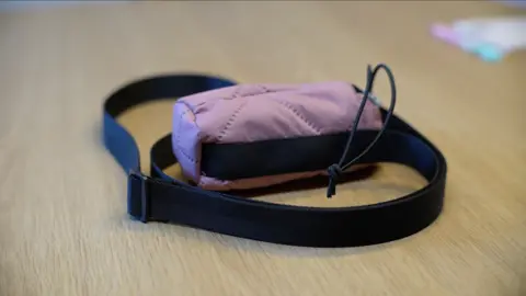 Jamie Niblock/ BBC The pouch is pink with a stitched quilted like design, and sits on a table. It has a black drawn string to ensure safe keeping of the ambulatory pumps, as well as a black adjustable strap.