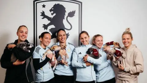 Birmingham Dogs Home Six women wearing sporting clothes are standing in a row in front of a large Aston Villa logo and they are each holding a puppy.