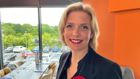 Woman with short blonde hair red lipstick black blazer stood infront of window 