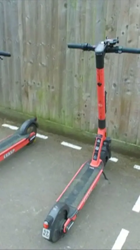 Orange and black e-scooter parked on pavement next to a wooden fence