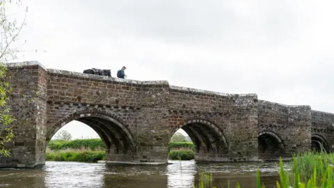 sesmlphotography Roger and Scarlet walking on Whitemill Bridge near Sturminster Marshall. It is cloudy.