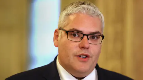 PA Media Gavin Robinson speaking. He has grey hair and black glasses. He is wearing a black suit blazer and white shirt.