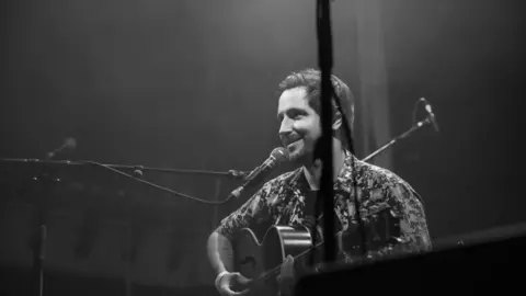 Aperturejackal Black and white image of Ollie Thomas sat on stage with his guitar, in front of a microphone, smiling. Ollie has short hair and dark stubble. He wears a t-shirt with a short-sleeved shirt worn over it with the buttons undone. A balcony is visible in the background, another microphone stand is in the foreground.