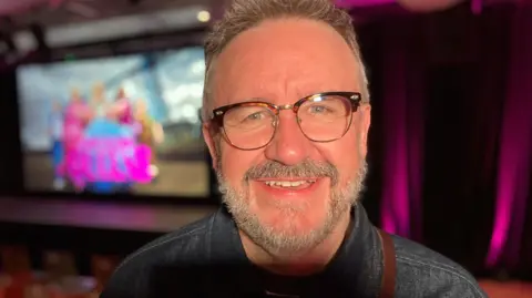 Mark Benton is smiling broadly and sitting down against a blurred but colourful poster of the show. He has short hair and a grey beard and is wearing glasses. 