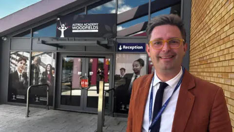 Image shows glass doors to a school building in the background. To the left of shot is a man wearing glasses, with a light brown suit on,a white shirt and black tie. 