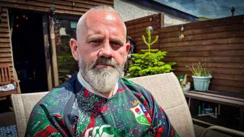 Jason sits in a garden chair on a patio. He has on a lions tour rugby jersey, short grey hair and a clipped grey beard. 
