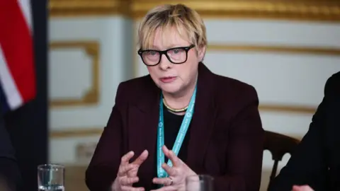 Getty Images Angela Eagle, who has short blonde hair and glasses, speaks while seated at a table. She is wearing a dark suit and blue lanyard, with a union flag in the background. 