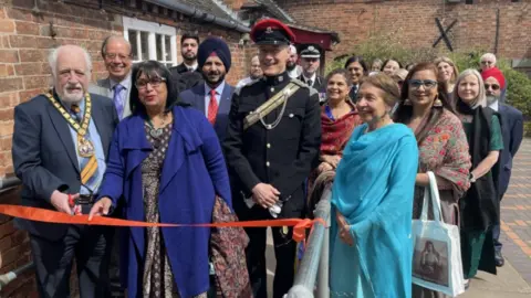 The chairman of Leicestershire County Council cuts the red ribbon, behind him are the Bosworth sikh women's group and other guests 