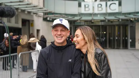 Getty Images Jamie Laing and Sophie Habboo outside BBC Broadcasting House in 2024. Jamie wears a white cap and black waterpoof while Sophie leans into him, wearing a black leather jacket. She has long blonde hair and smiles at a camera.