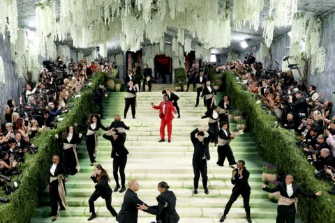 MG26/Getty Images para el Museo Metropolitano/Vogue. Joshua Henry actúa durante la Gala del Met 2026, dedicada al arte del vestuario, en el Museo Metropolitano de Arte el 4 de mayo de 2026 en Nueva York. Henry luce un esmoquin rojo cruzado con adornos plateados en las solapas. Se sitúa en las escaleras y canta mientras actúa para la prensa.