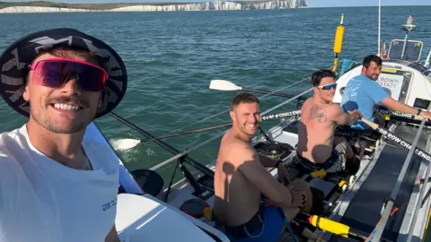 Rowmads A selfie taken by a man wearing a white top, sunglasses and a hat. Behind him are three men sitting and rowing, but turning around to smile at the camera. Two are shirtless and the one sitting towards the front is wearing a blue top.