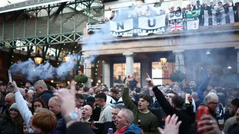 A crowd of Newcastle United fans singing with their hands in the air in Covent Garden. Someone is holding a flare.