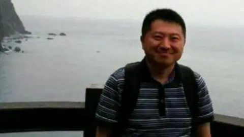 X/Liu Hu Liu Hu wearing a blue polo shirt and smiling as he poses for a photo. Behind him is the sea.