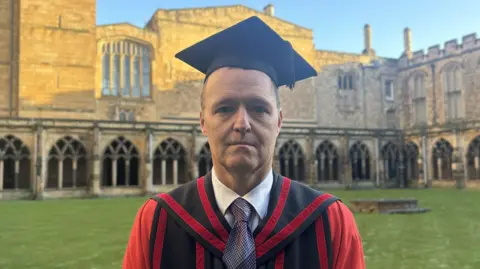 BBC News A man wearing a red and black cloak with a black graduation hat, standing in front of a cathedral. 