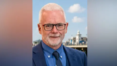 Michael O'Keeffe está sonriendo. Lleva gafas y al fondo se ve el muelle de Clacton.