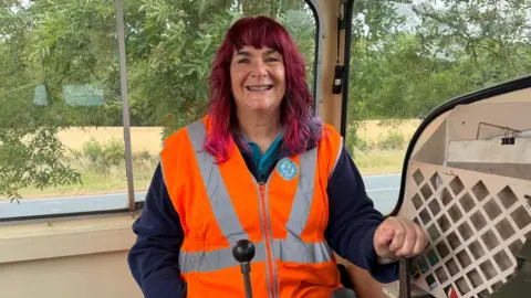 Katy has her hand on the break and is smiling. She is also in an orange high vis vest and is in the locomotive.