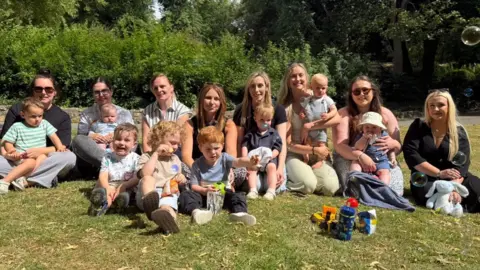 Eight women sitting in a park and smiling at the camera on a sunny day. In front of them are eight boys who are sitting down on the grass or on their mum's knee. Some are eating, others are looking at a bubble machine in the distance. Children's toys can be seen.