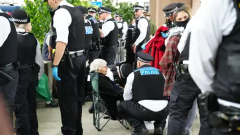 PA/Jeff Moore Reverend Sue Parfitt sitting in a green camping chair, speaking wit an officer who is crouched down beside her. They are surrounded by more police officers wearing black and white uniforms and stab proof vests.