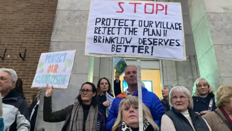 Men and women protesters stand on the steps of the town hall with placards. One says "protect our countryside" and another reads "Stop! Protect our village reject the plan!! Brinklow deserves better!"