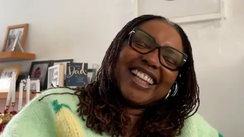 Yvonne Johnson laughing sat in her living room in her wheelchair, with a portrait of her lips behind her.