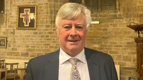 Nadia Lincoln LDRS Graham Ferrie, who has short, white hair and is wearing a black blazer, white shirt and a multi-coloured tie. The picture has been taken in a church. Chairs, a brick wall and part of the pulpit are in the background.