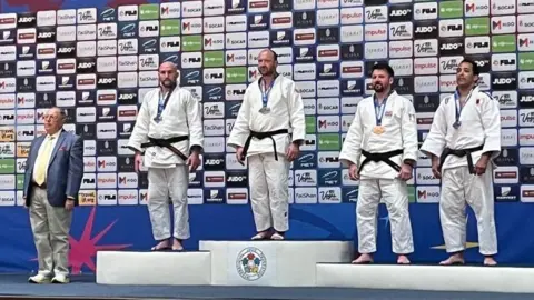 Judo Veterans World Champion Shaun Mitty standing on the podium after winning gold.
He is wearing his judo uniform alongside three other men who are standing on the silver and bronze podiums.