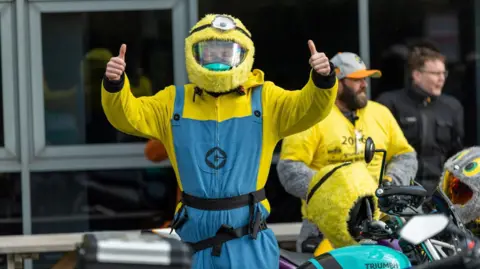 A person dressed in a yellow and blue fancy dress costume. The outfit includes a yellow furry helmet with a single large eye on to and a blue and yellow jumpsuit.  The person is giving two thumbs up. Several motorcycles, including one with a Triumph logo, are parked around them.