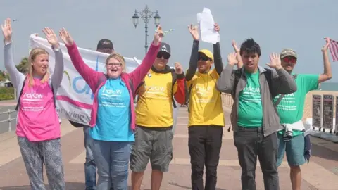 Halow People wearing brightly coloured Halow t-shirts walk along a seafront promenade holding a large flag, with railings and the sea in the background.