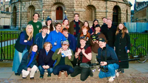 Max Morgan Some of the cast and crew of a film on set. They are standing and squatting in a group and smiling at the camera. Several of them are holding film equipment.
