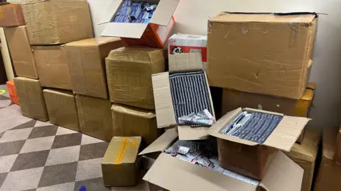 A dozen cardboard boxes are stacked up against a white wall. Some are open, with the contents appearing to be white tablets in navy film. 