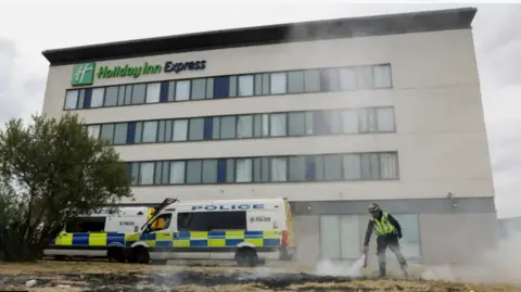 The Holiday Inn at Manvers, Rotherham,  with two police vans parked in front of it. A man in a high vis jackets is using a fire extinguisher on scorched grass in front of the building.