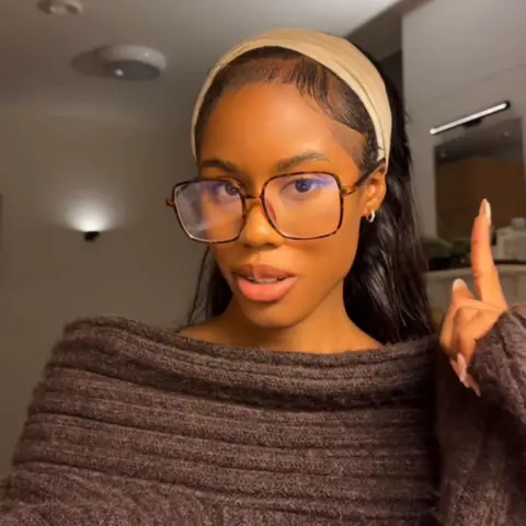 @ayoolaib A selfie of Ayoola wearing a brown off the shoulder knitted jumper and pointing to a wall light behind her