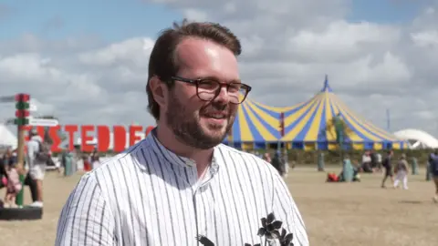 A man with a white shirt and gray stripes is stood on a dry grassy field. He has brown hair and is wearing black glasses, in the background is a sign for the Eisteddfod and a large yellow and blue striped tent. There is also a blue sky predominately covered by white fluffy clouds.