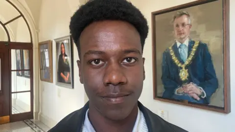 A man standing in a corridor, he has black hair and is wearing a black jacket with a white shirt