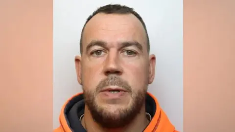 Daniel looks at the camera with a blank expression. He wears a bright orange hoodie and has a dark beard and thinning short dark hair.